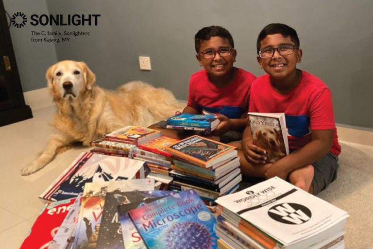 two brothers sit behind a pile of homeschool curriculum. A brown dog sits beside them.